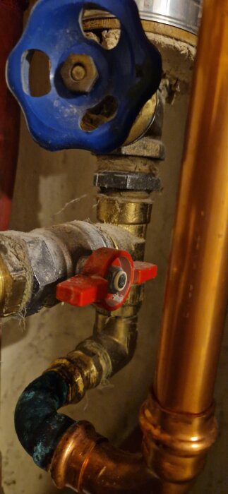 Ventil och kopparrör med blått vred i en vatteninstallation, omgivna av mässingsdelar och oxidbildning på röret.