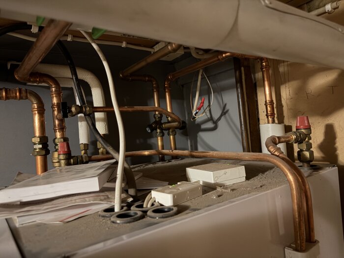 Värmesystem med kopparrör, ventiler och elektronik i ett dammigt område, rören har röda toppar och flera kopplingar synliga.