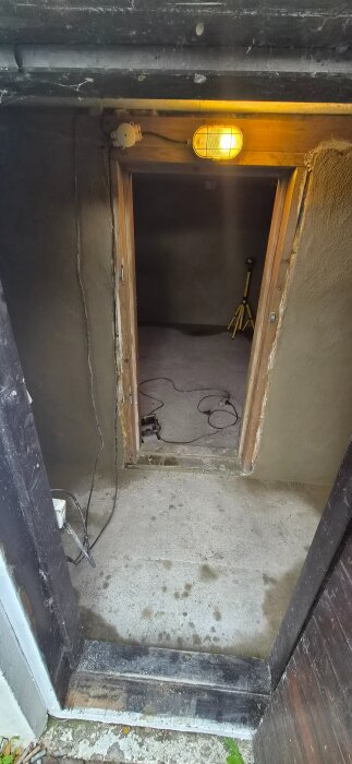 En gråputsad dörröppning med en tänd taklampa och elkablar på betonggolvet, förberedelser för byggprojekt.