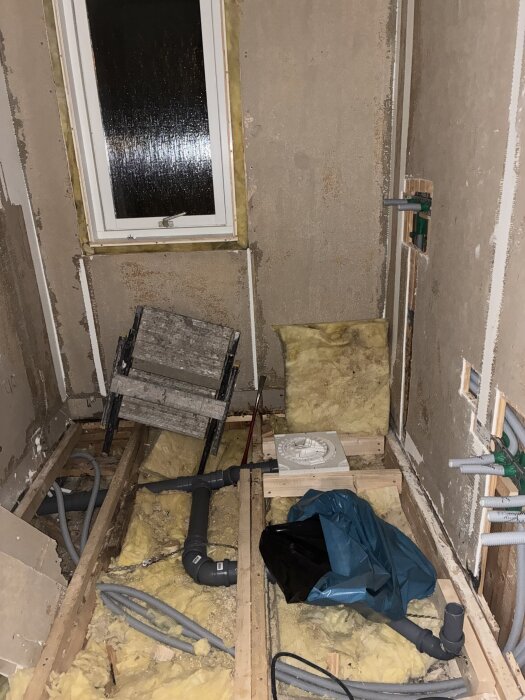 Renovering av rum med öppna golvbjälkar, isolering och rörledningar, samt en stege och fönster.