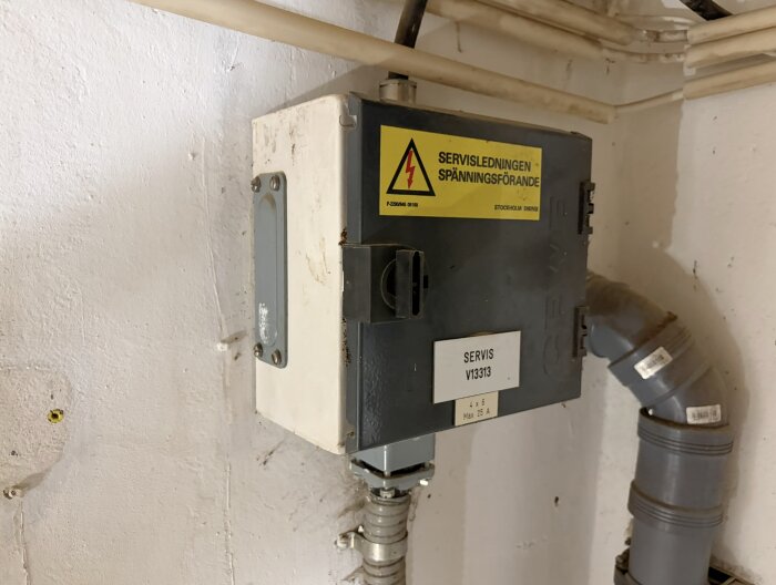 Grå elcentral med varningsskylt för spänning, placerad på en vägg tillsammans med rörinstallationer.