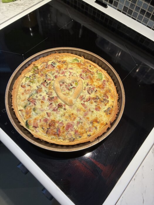 Paj med broccoli, purjolök, rostade solroskärnor och skinka i en rund form, toppad med ett degformat hjärta, står på en spishäll.
