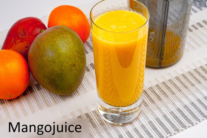 Glas med mangoshake bredvid apelsiner och mango på en randig duk, med texten "Mangojuice".