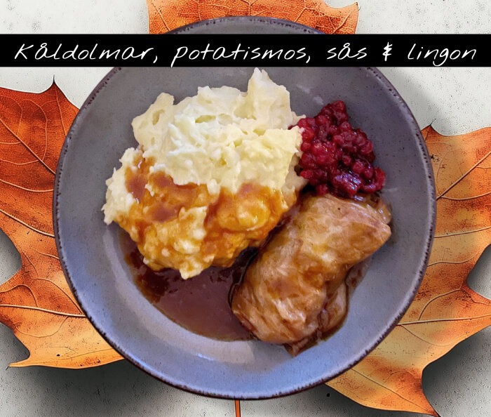 Tallrik med kåldolmar, potatismos, brun sås och lingonsylt, dekorerad med höstlöv i bakgrunden.