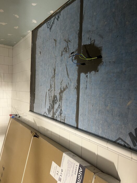 Väggen i badrummet med halvkaklat område, tätskikt på övre delen och eluttag exponerat. Kartong och verktyg syns nedanför väggen.