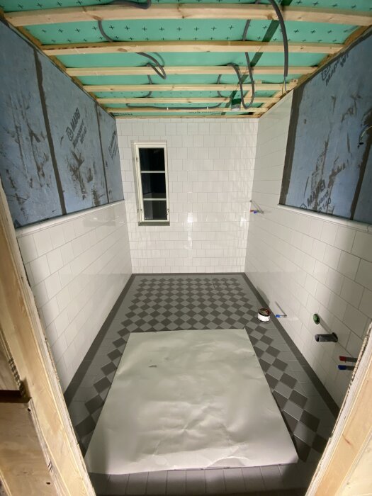 Halvkaklat badrum under renovering med bröstning och grön isolering i taket. Golvet har ett rutigt mönster och ett papper täcker en del av golvet.