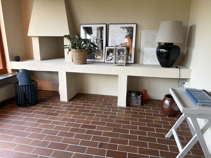 Vardagsrum med eldstad, krukväxt och bilder på en vit hylla samt rödbrunt klinkergolv.