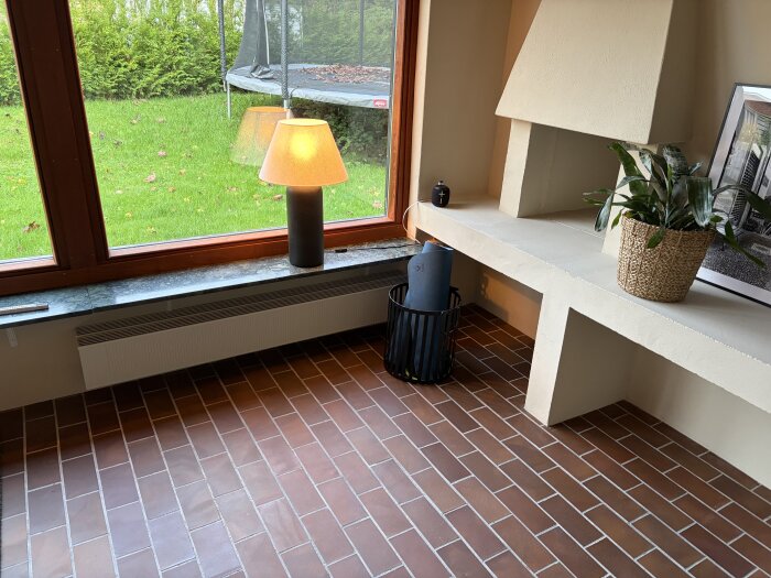 Rum med klinkergolv, ett stort fönster med utsikt mot en gräsmatta med studsmatta, en bordslampa och en krukväxt på en bred fönsterbänk.