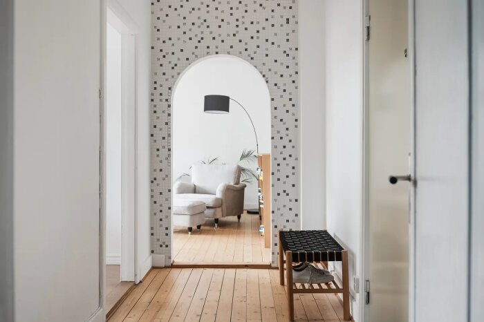 Korridor med trägolv, vägg med mosaikdetaljer och bågformad öppning mot vardagsrum med fåtölj och golvlampa, skohylla i förgrunden.