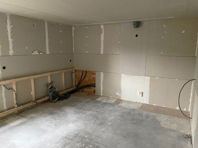 Tomt rum under renovering med osb bakom gipsskivor, synliga kablar och installationer i nedre hörnet.