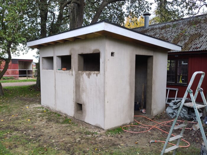 Nybyggt putsat uthus med nischer, omgivet av träd. Stege, sladd och material syns runt byggnaden.