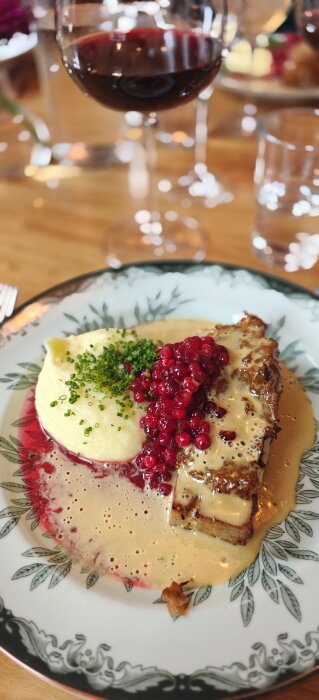Kålpudding på tallrik med potatismos, lingon och gräddsås, glas rödvin i bakgrunden.