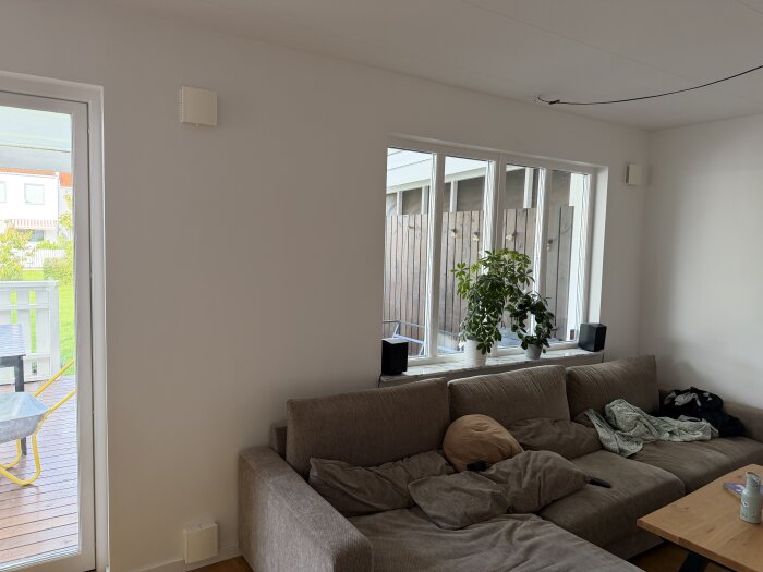 Soffhörna med en grå soffa täckt av kuddar och filtar, ett träbord och en grön växt i fönstret, med utsikt mot en terrass och trädgård.