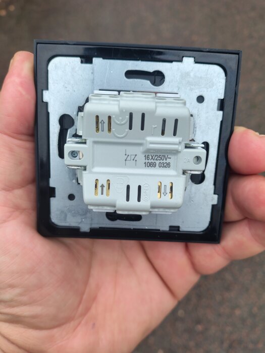 Malmberg strömbrytare sedd bakifrån, hålls i en hand utomhus. Visar anslutningspunkter och märkning, inklusive 16A, 250V symboler och skruvhål.