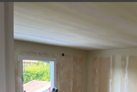 Rum under renovering med nyligen spacklade väggar och ett fönster som vetter ut mot en trädgård och röda tak.