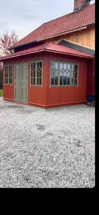 Röd byggnadsfasad med gröna dörrar och fönster, valmat tak med klickfals, och ett grusbelagt område framför.
