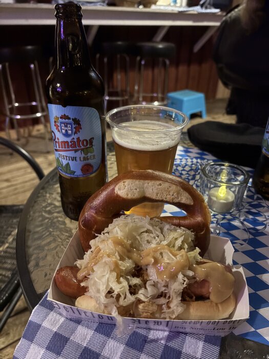 Flaska Primator lager, plastglas med öl, och en bratwurst med surkål och senap i bröd, dekorerat med en pretzel.