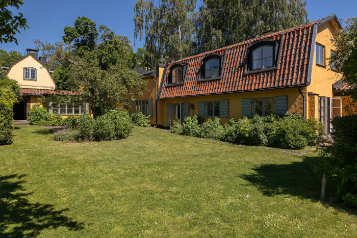 Gult hus på Djurgården från 1794 med tegeltak och stora fönster, omgivet av lummig trädgård och gräsmatta.
