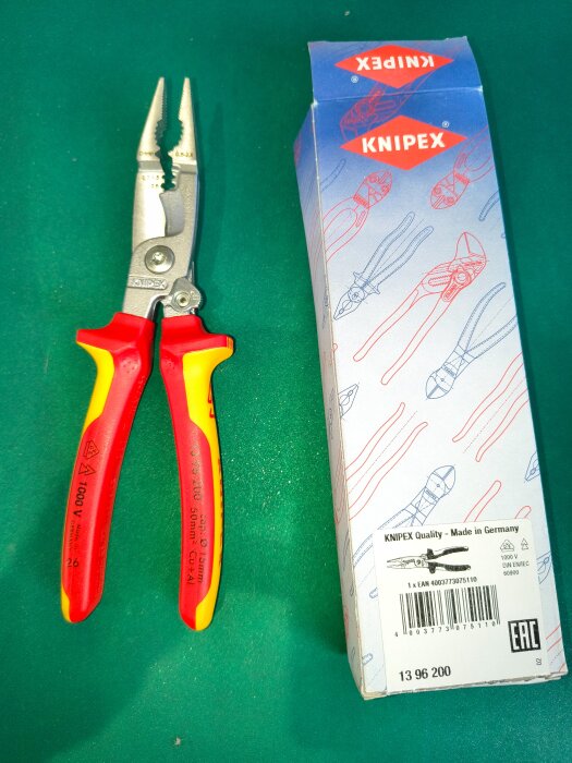 Röd-gul kombinationstång från Knipex bredvid sin förpackning på en grön yta.