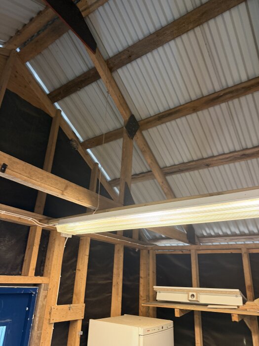 Invändigt i ett garage utan undertak, visar synliga takstolar och takplåt. En lysrörslampa hänger ner från taket.