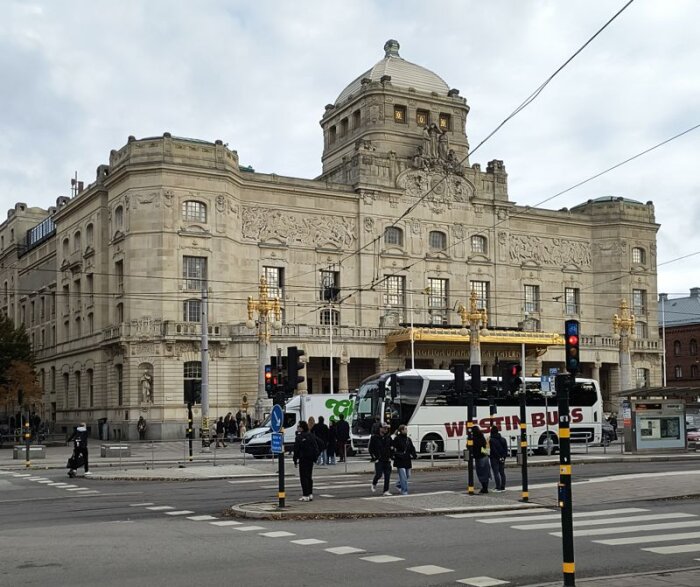 Ståtlig byggnad i klassisk stil med detaljerade fasadutsmyckningar, i förgrunden syns en buss och fotgängare vid ett övergångsställe.