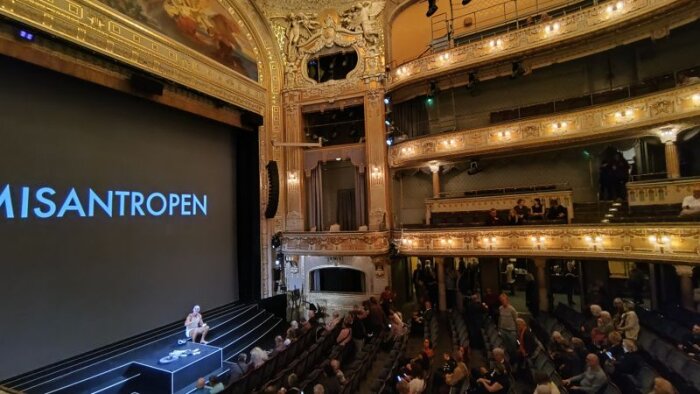 Interiör av teater, scen med texten "Misantropen", publik i salongen, dekorerade väggar och balkonger.