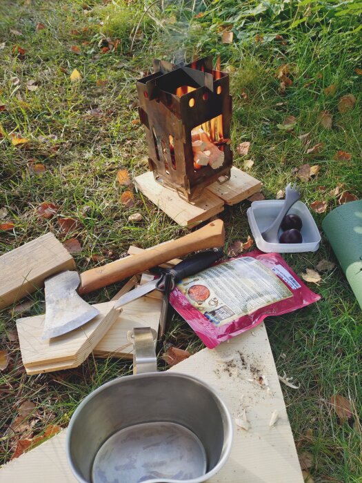Eldstad av metall utomhus i gräs, med yxa, kniv, metallkopp och matförpackning runtom.
