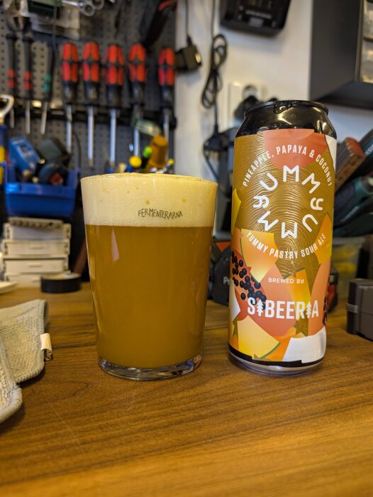 Ett glas öl står på ett träbord bredvid en ölburk med texten "Pineapple, Papaya & Coconut Yummy Pastry Sour Ale" i en verkstadsmiljö.