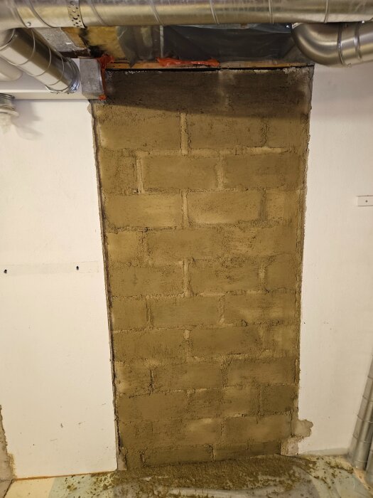 Betonghålstenar med löst slammat c-putsbruk applicerat med kalkborste, omgivna av rör och vit vägg.