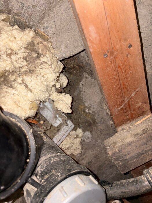 Isolerad öppning med fogskum i källare, del av träpanel borttagen, svart avloppsrör synligt.