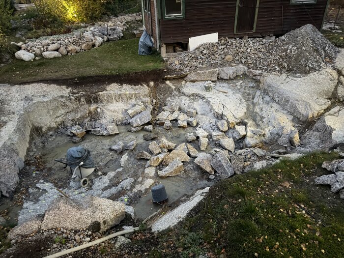 Stenbrott med uppbrutna stenar nära ett trähus och gräsområde, med en borrmaskin och diverse verktyg på platsen.