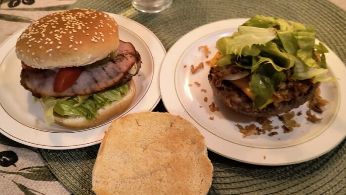 Två hamburgare på tallrikar, en med skivat kött, tomat och sallad i bröd, och en utan bröd, med smält ost, sallad och stekt lök.