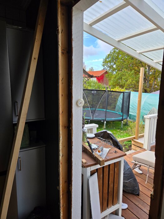 Ingångsparti under renovering med avlägsnad isolering och tillfälligt uppställt byggmaterial, utsikt mot altan och trädgård med studsmatta.