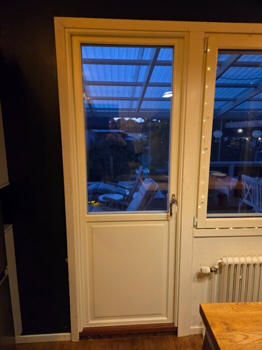 Vit ytterdörr med glasruta och synligt dörrfoder vätter mot en inglasad altan. Till höger om dörren finns ett element och ett bord.