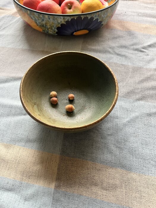 Grön skål med fyra hasselnötter på randig duk; i bakgrunden en färgglad fruktskål med äpplen och citroner.
