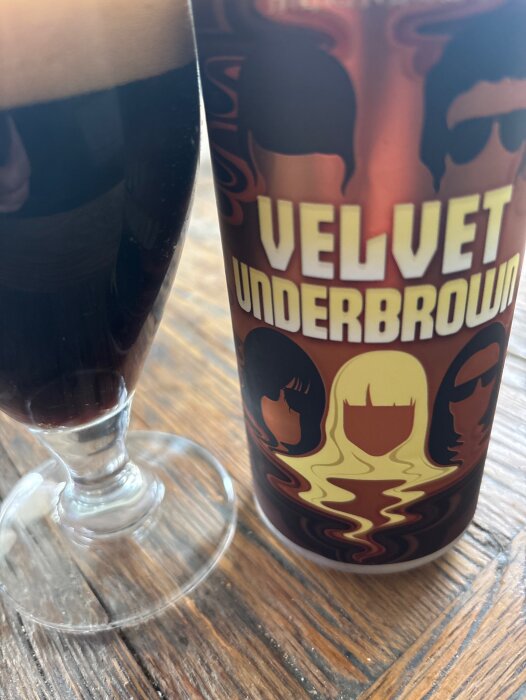 En glas Brown Ale bredvid en burk med texten "Velvet Underbrown", stående på ett träbord.