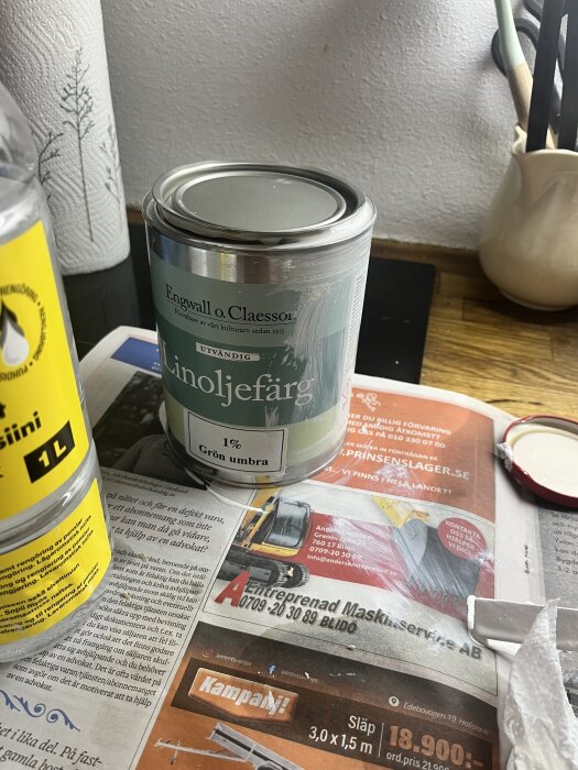 Burk med vit linoljefärg med 1% grön umbra från Engwall o. Claesson, står på tidning på ett bord, bredvid en flaska med gul etikett.
