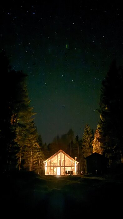 Upplyst stuga om natten i skogen, med stjärnhimmel ovan.