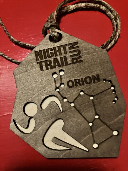 Brun medaljong med texten "Night Trail Run" och "Orion" samt motiv av löpare och stjärnbild, hängande i ett snöre på röd bakgrund.