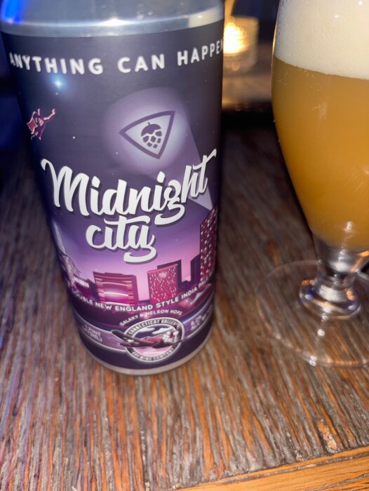 Burk med Midnight City, New England style IPA, bredvid ett glas fyllt med öl, på ett träbord.