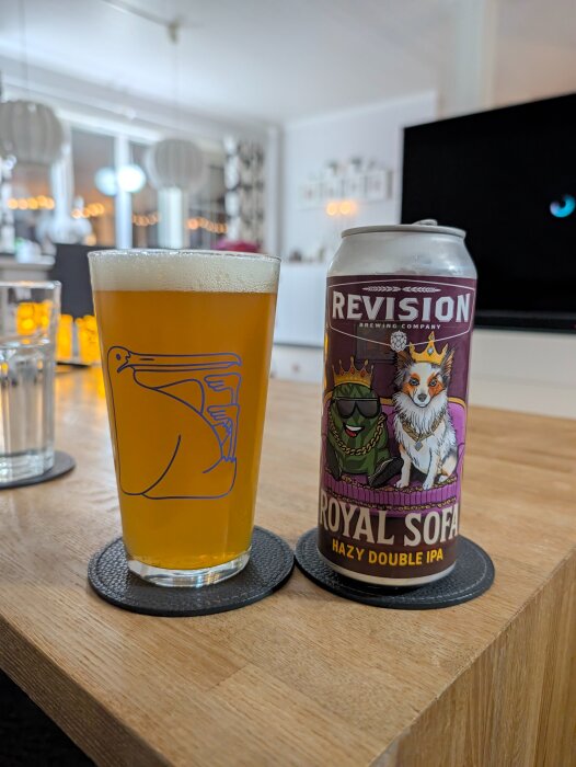 Glas med gyllene öl och en burk Revision Royal Sofa Hazy Double IPA på ett bord i ett vardagsrum.