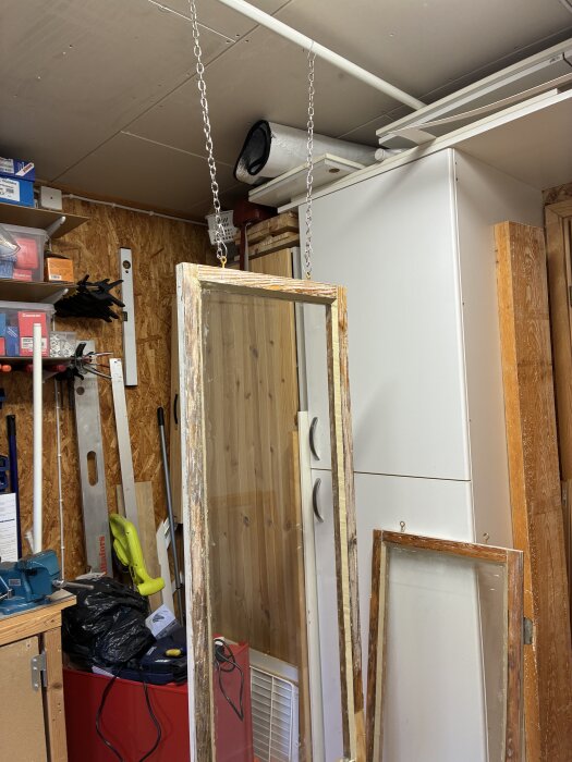 Fönsterbåge upphängd med kedjor inomhus för målning i ett förråd med verktyg och förvaringsutrustning.