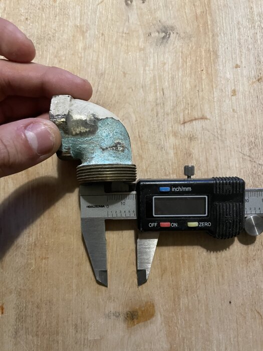 En person håller ett korroderat radiatorböj jämte en digital skjutmått för att mäta dimensionerna, på en träyta.
