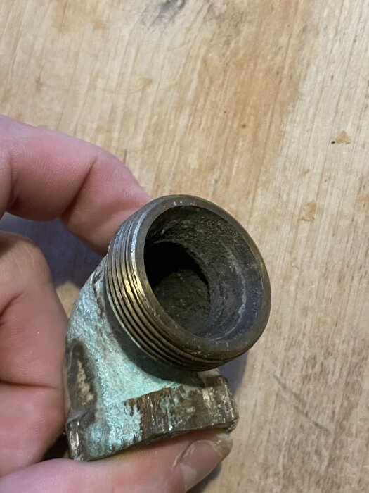 Radiatorböj i metall med spricka, hålls av en hand, träbakgrund synlig.