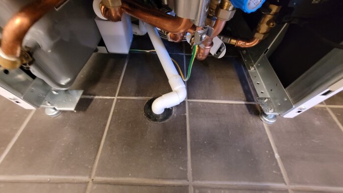 Rörinstallation med plaströr och kopparrör under en värmepump, med ett plaströr som leder till ett avloppshål på golvet.