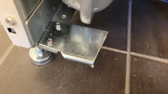 Bild av en metallskena under en värmepumpsinstallation på golvet, med synliga nitar och stående på kakelplattor.