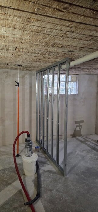 Källarutrymme under renovering med synligt planktak, metallstomme för vägg och rördragning. Bucket och verktyg synliga på golvet.