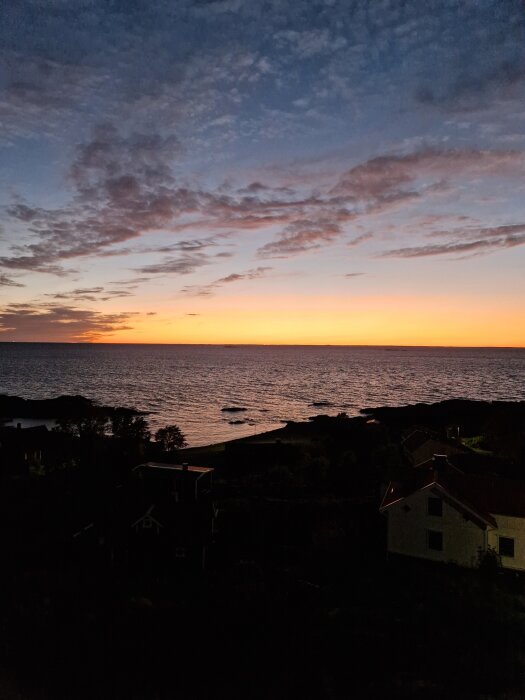 Solnedgång över havet sett från land, med silhuetter av träd och hus i förgrunden under en färgstark himmel.