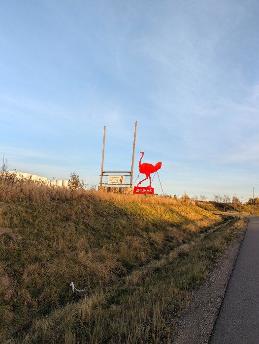 Röd hästfigur placerad på en grässlänt bredvid en väg, under en klar blå himmel.