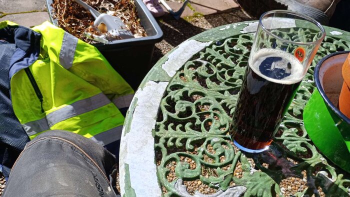 Grön metallbord med ett glas mörk öl, en reflexjacka och en behållare med löv och skräp i bakgrunden på en uteplats.